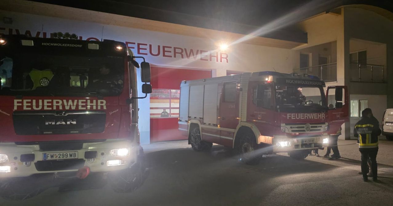 Zwei rote Feuerwehrfahrzeuge sind nachts vor einer Feuerwache geparkt. Das Gebäude hat ein Schild mit der Aufschrift 'FEUERWEHR'.