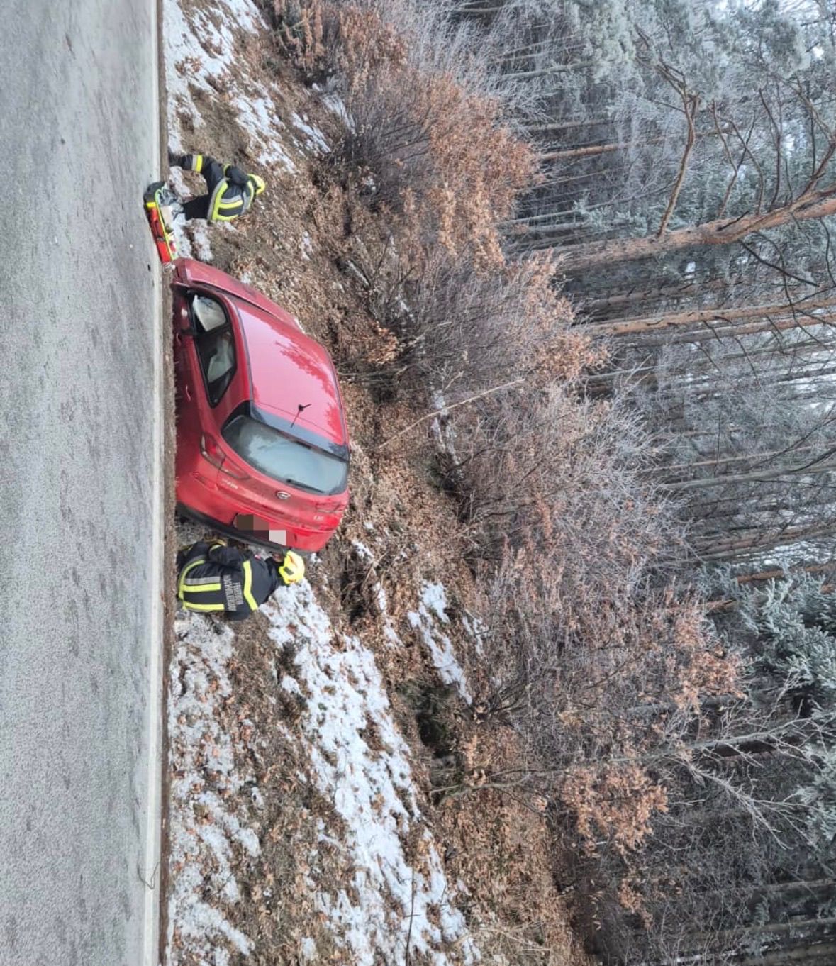 Zwei Feuerwehrleute befinden sich am Straßenrand, einer in der Nähe eines roten Autos. Das Auto steht am Straßenrand, möglicherweise nach einem Schleudern. Das Gebiet ist verschneit mit Bäumen.