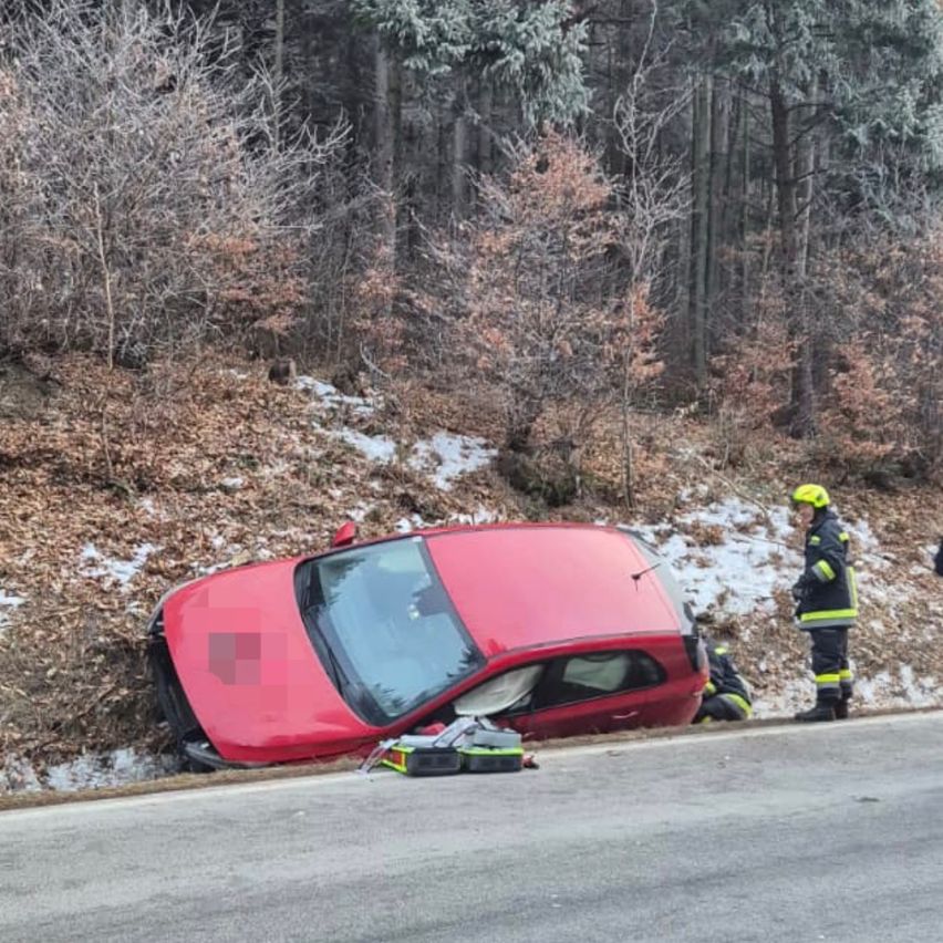 Ein rotes Auto liegt auf der Seite am Straßenrand. Feuerwehrleute kümmern sich um die Szene, einer steht in der Nähe des Autos.