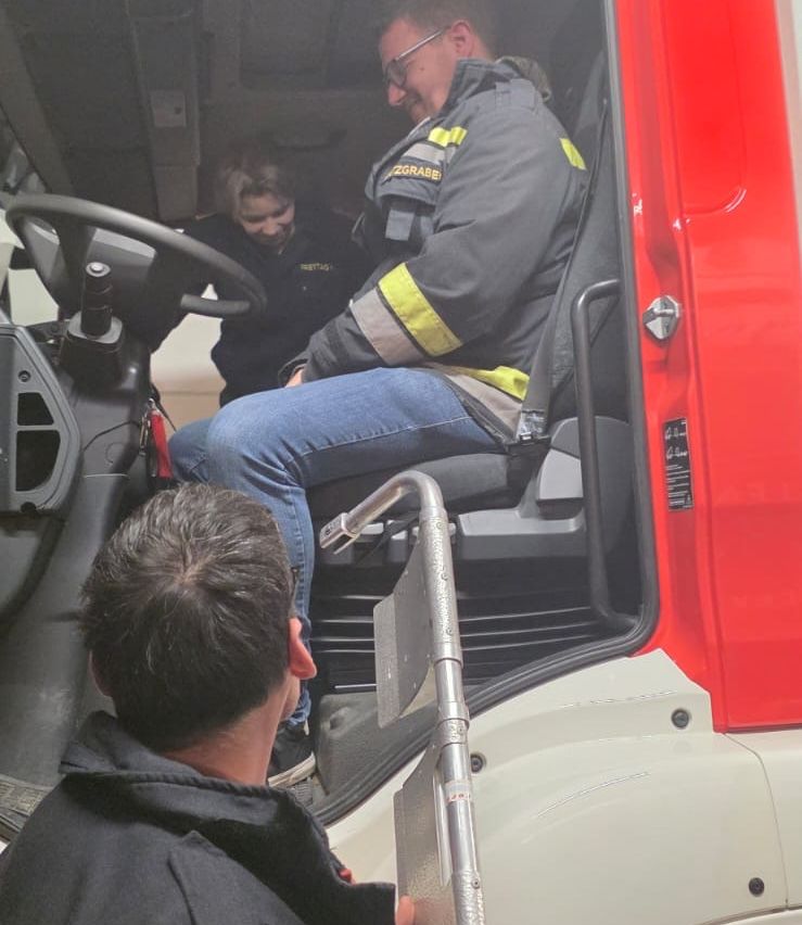 Ein Feuerwehrmann sitzt auf dem Fahrersitz eines roten Feuerwehrwagens, während eine andere Person neben ihm steht und möglicherweise etwas erklärt. Eine dritte Person schaut ihnen von vorne zu.