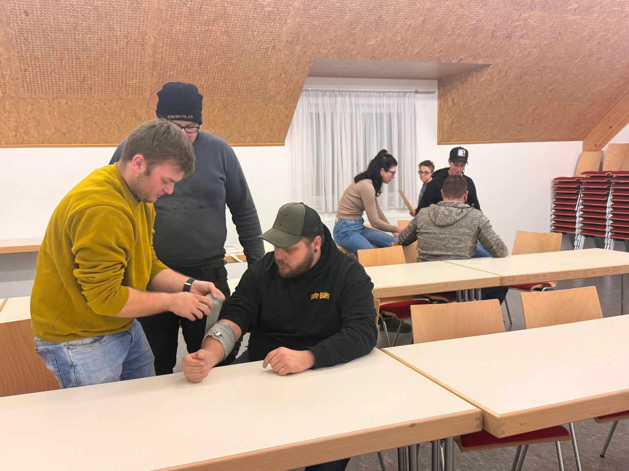 Eine Gruppe von Menschen versammelt sich in einem Raum, wahrscheinlich für eine Schulung. Ein Mann sitzt auf einem Stuhl, und ein anderer Mann steht neben ihm und fixiert seinen Arm. Einige Leute stehen hinter ihnen, und einige sitzen auf den Stühlen. Ein Tisch steht vor ihnen, und ein Fenster befindet sich dahinter.