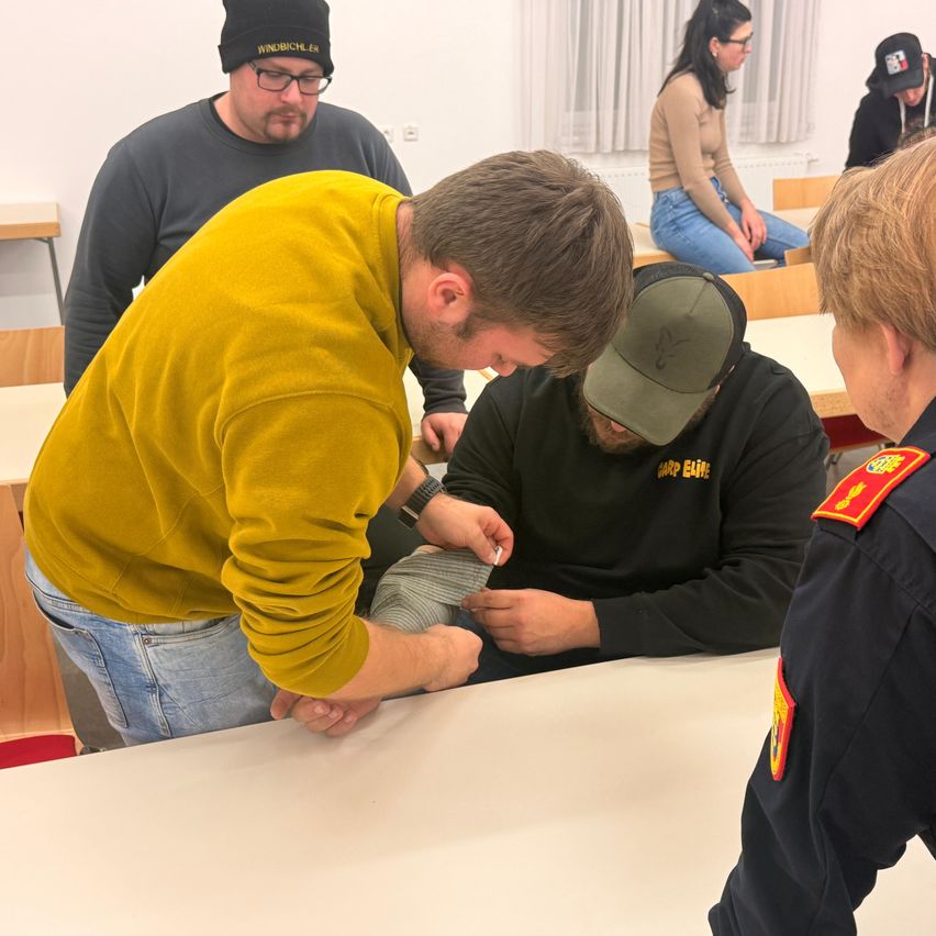 Mehrere Personen sind drinnen; ein Mann beugt sich über einen sitzenden Mann und überprüft möglicherweise seine Schuhe. Ein weiterer Mann mit Hut und eine andere Person beobachten. Eine Frau in Jeans sitzt hinter ihnen.