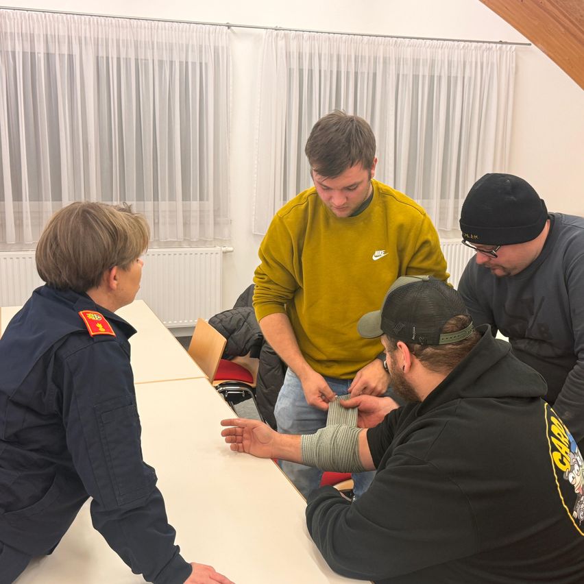 Ein Mann mit einem bandagierten Arm wird von einem anderen Mann in einem Besprechungsraum geholfen. Ein Ausbilder in Uniform beobachtet sie. Hinter ihnen befinden sich Fenster mit weißen Vorhängen und einem Heizkörper.