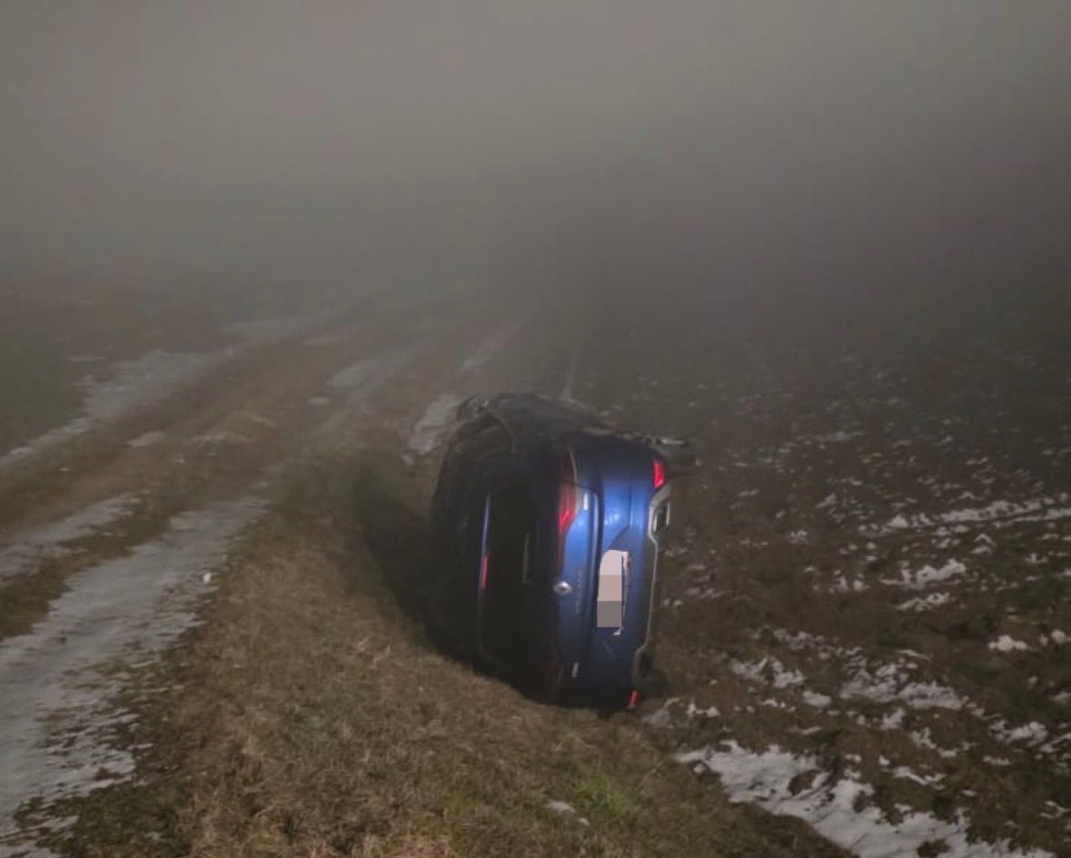 Ein blaues Auto liegt auf der Seite auf einem Grasfeld mit Schneeflecken. Die Szene ist neblig, und der Himmel ist bedeckt.