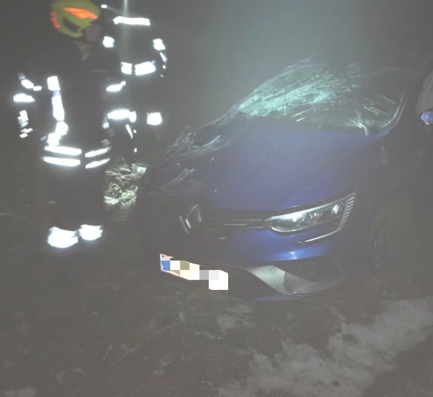 Zwei Feuerwehrleute stehen neben einem demolierten blauen Auto, einer trägt einen Helm. Die Front des Autos ist stark beschädigt und das Nummernschild ist unkenntlich gemacht.
