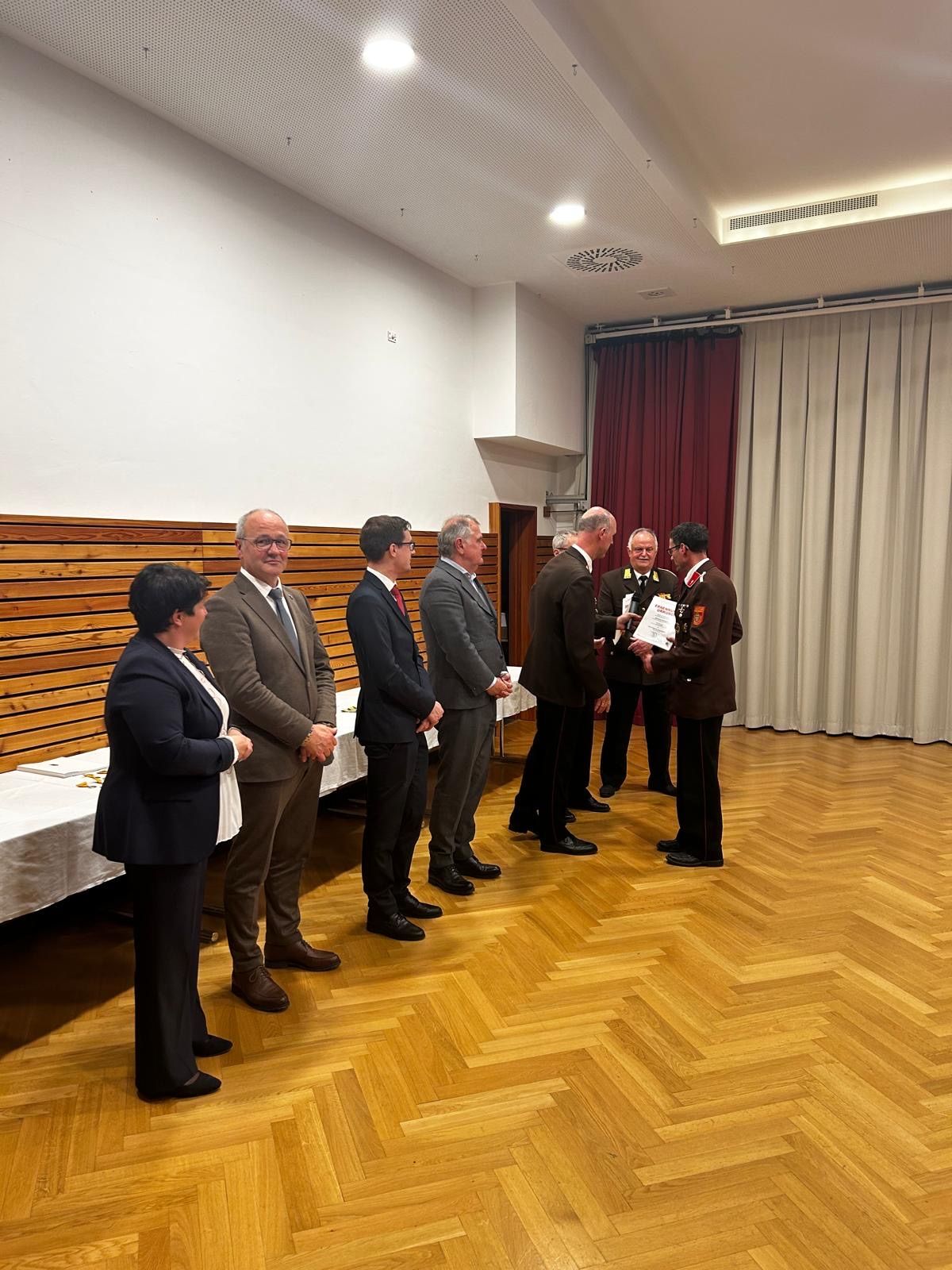 A group of people in formal attire stand in a room, possibly during a ceremony. They are arranged in a line with one person holding a paper.