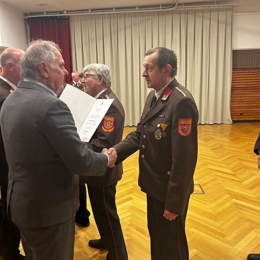Two men in suits shake hands in a formal setting. One man wears a military uniform with medals and badges. They stand in front of a white curtain and wooden wall.