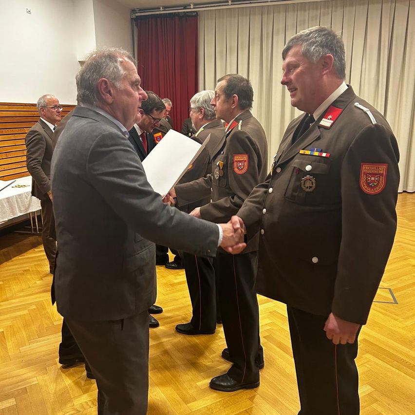 Two men in suits and uniforms are shaking hands in a formal event. Behind them are other men in suits and a table with a tablecloth.