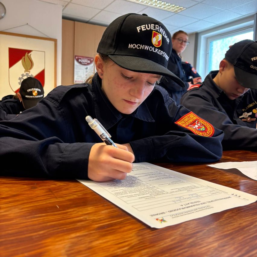 A woman in a uniform is filling out a document at a desk with several others wearing hats.