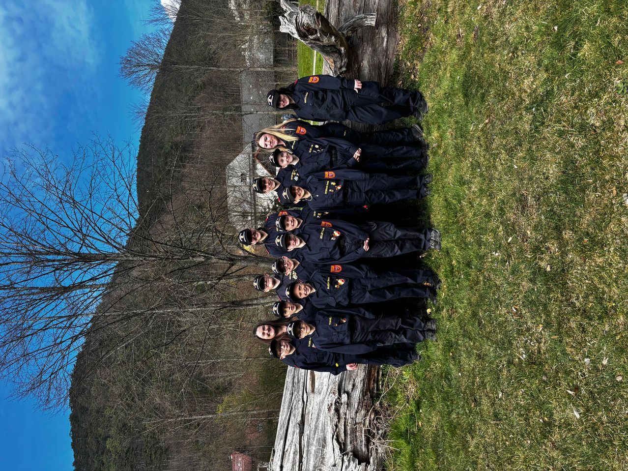 A group of firefighters in uniform stand on a grassy field, with a mountain and trees in the background.