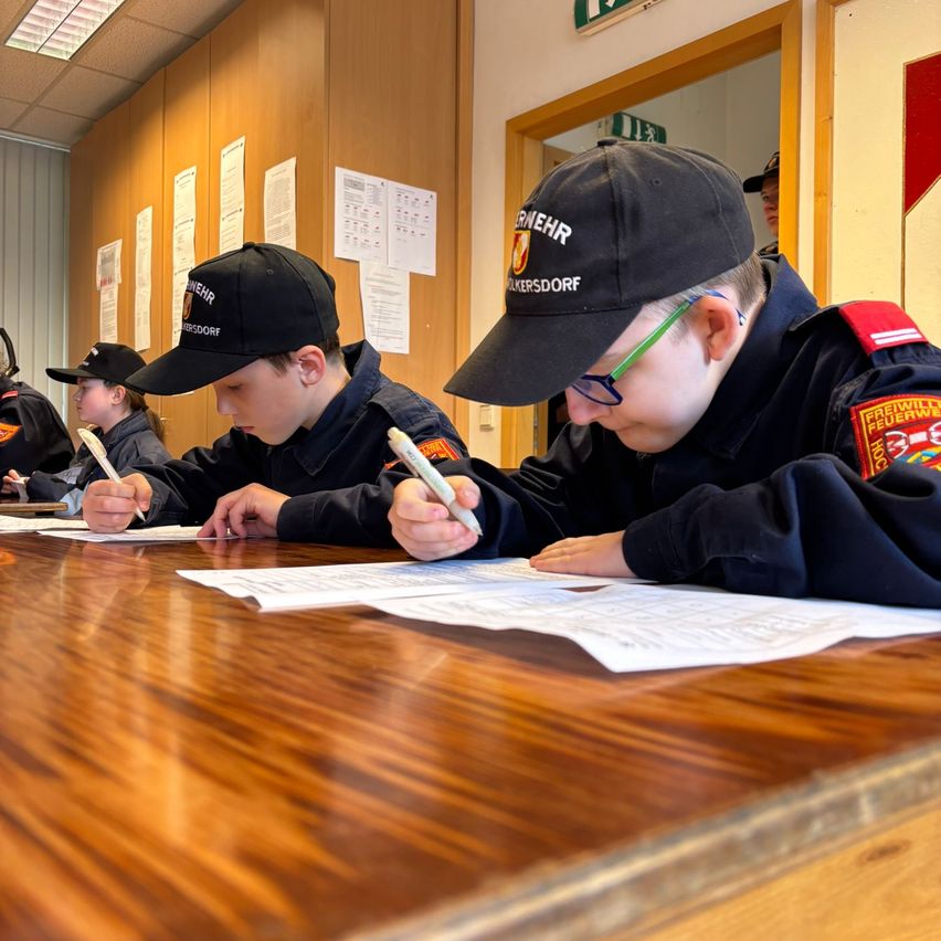 A group of children in uniform are sitting at a table and writing on paper with pens. They are wearing hats and glasses. The table is wooden.