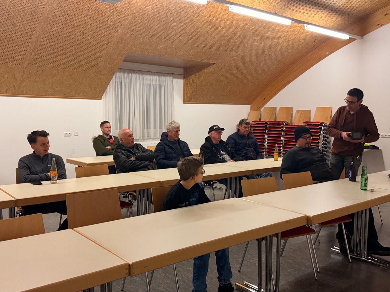 Eine Gruppe von Männern und einem Kind sitzen um Tische in einem Raum mit Holzwänden und weißen Vorhängen. Flaschen und andere Gegenstände stehen auf den Tischen.