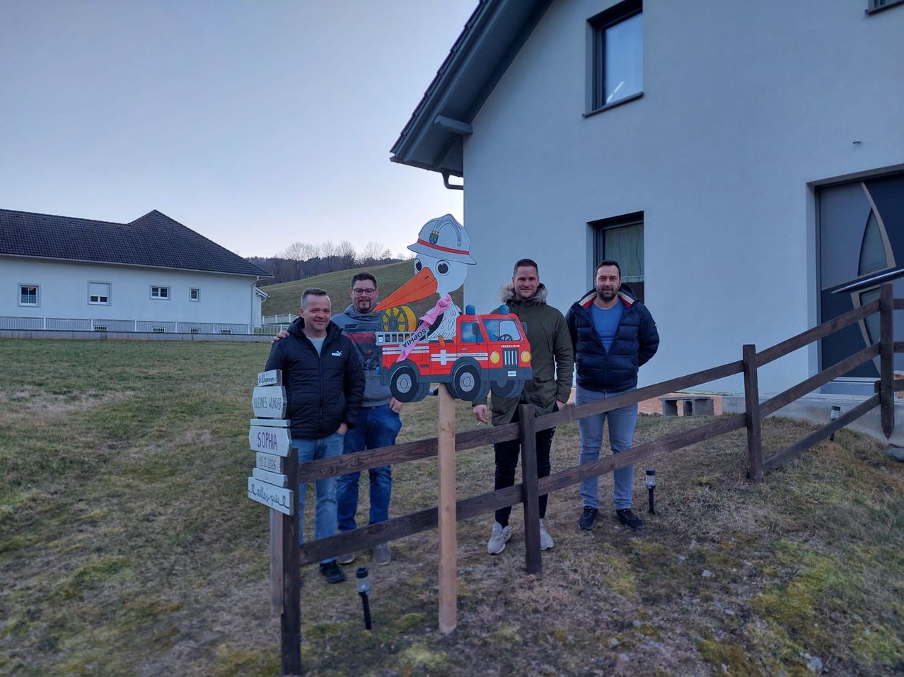 Four men stand in front of a house with a wooden fence. One of the men holds a cutout of a firefighter's hat.