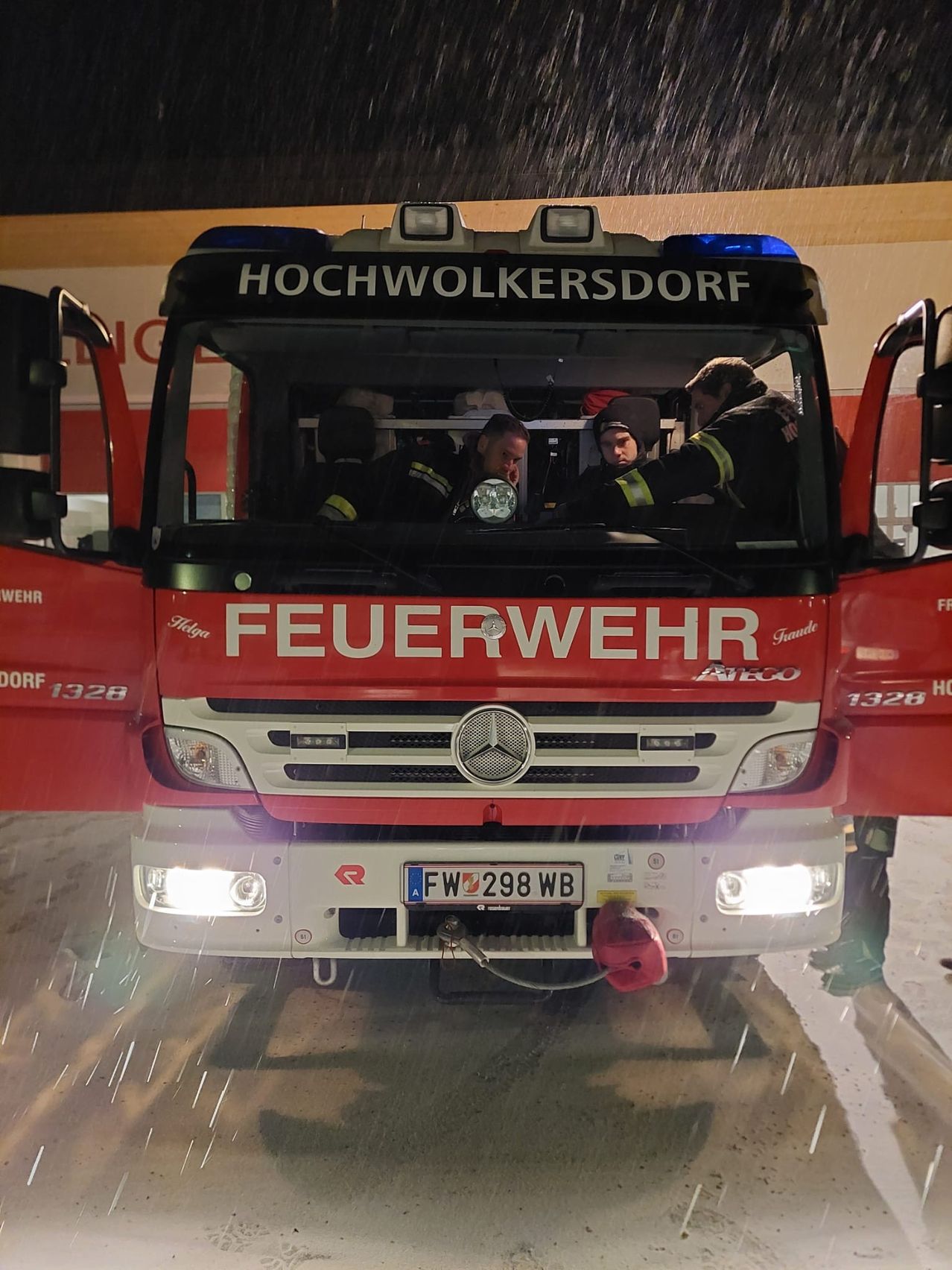 A red fire truck from Hochwolkersdorf, labeled with the number 1328, is parked in the rain. The front doors are open, and firefighters are seated inside.