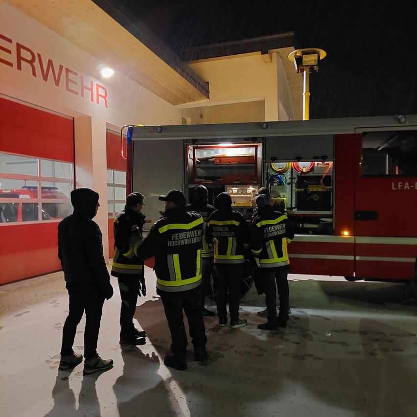 Firefighters are standing in front of a fire station at night, discussing something with one another.