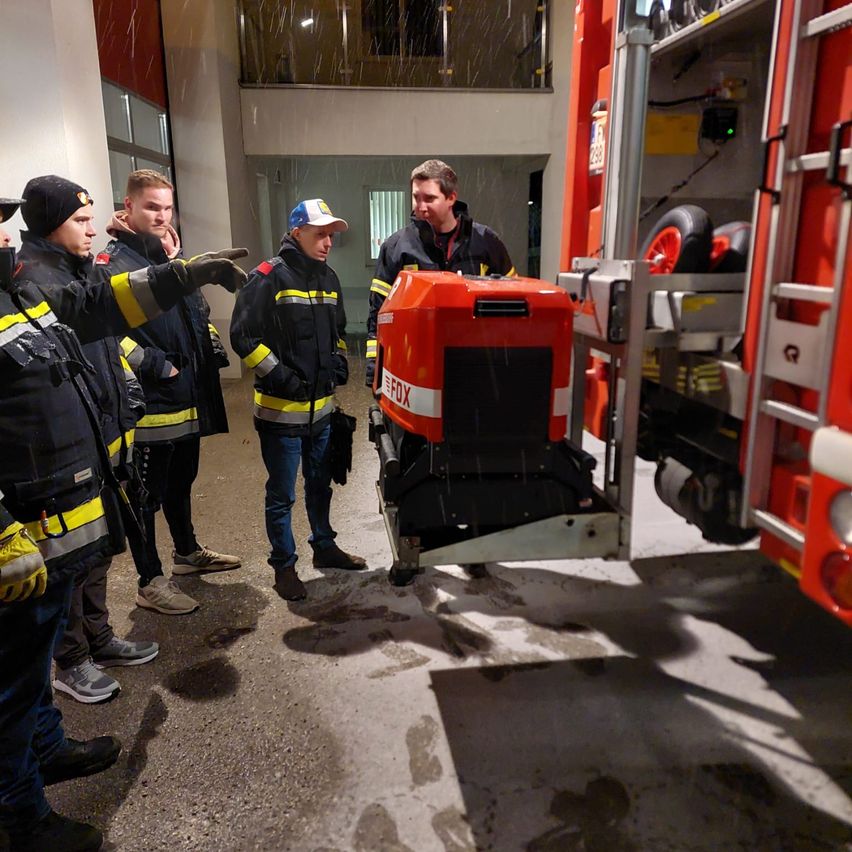 A group of firefighters is gathered around a fire truck, with one pointing at it. They are dressed in uniforms, hats, and gloves. The fire truck has an open door and visible equipment.