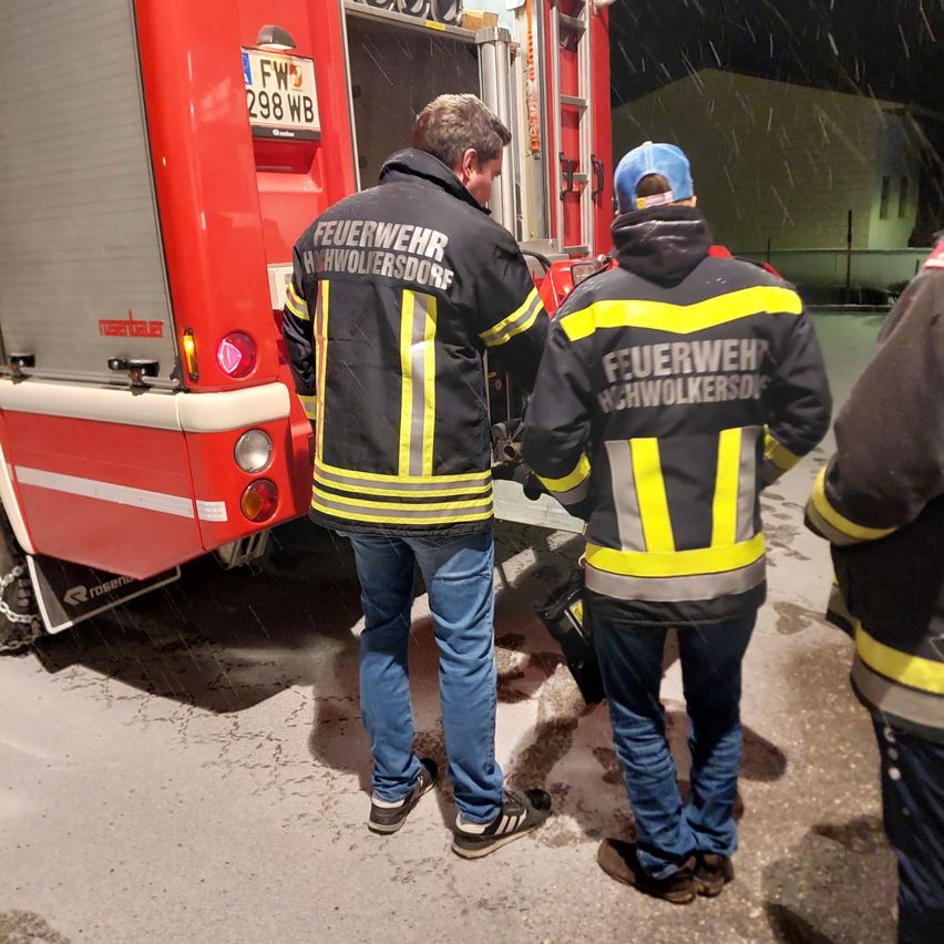 Two firefighters in protective gear are standing in front of a fire truck. It is night, and snow is falling. The fire truck has the license plate FW 298 WB.