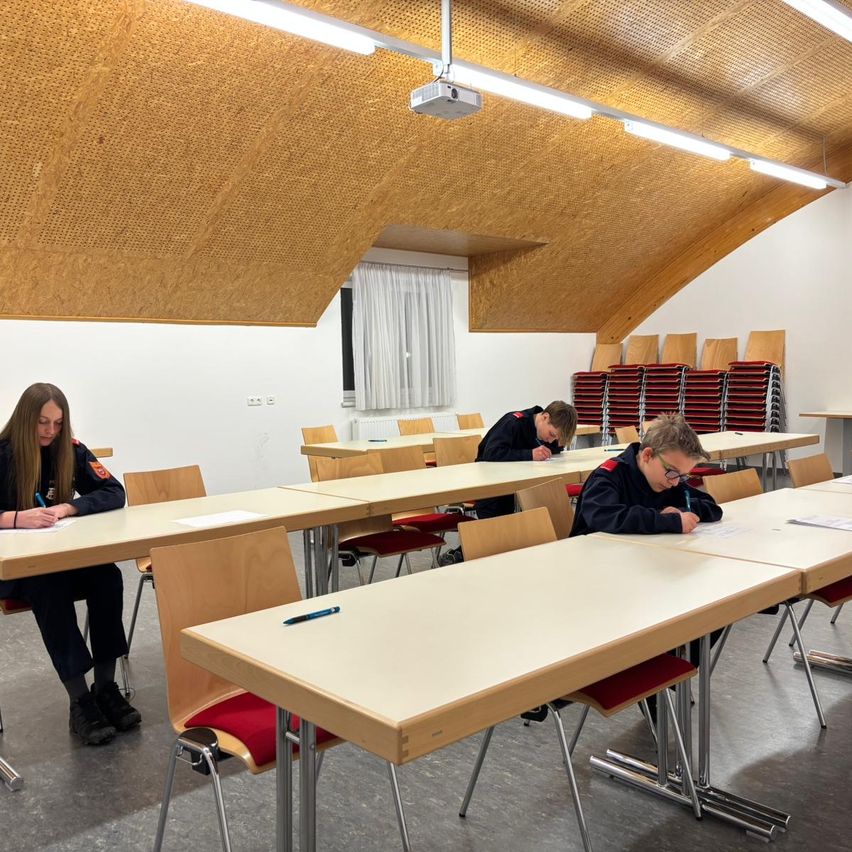 Three students are writing in a classroom with a curved ceiling and white walls. Each student is seated at a desk with papers and pens.