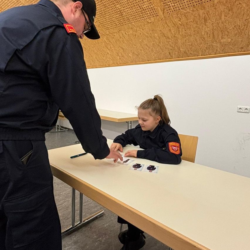 A uniformed person is teaching a young girl sitting at a desk. They are pointing at cards on the table. The room has a wooden wall and tiled floor.