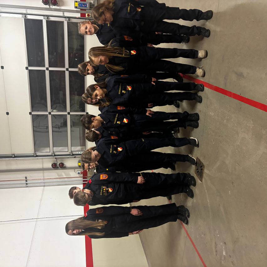 A group of firefighters in black uniforms with red logos stands in a row inside a fire station. They are facing away from the camera.