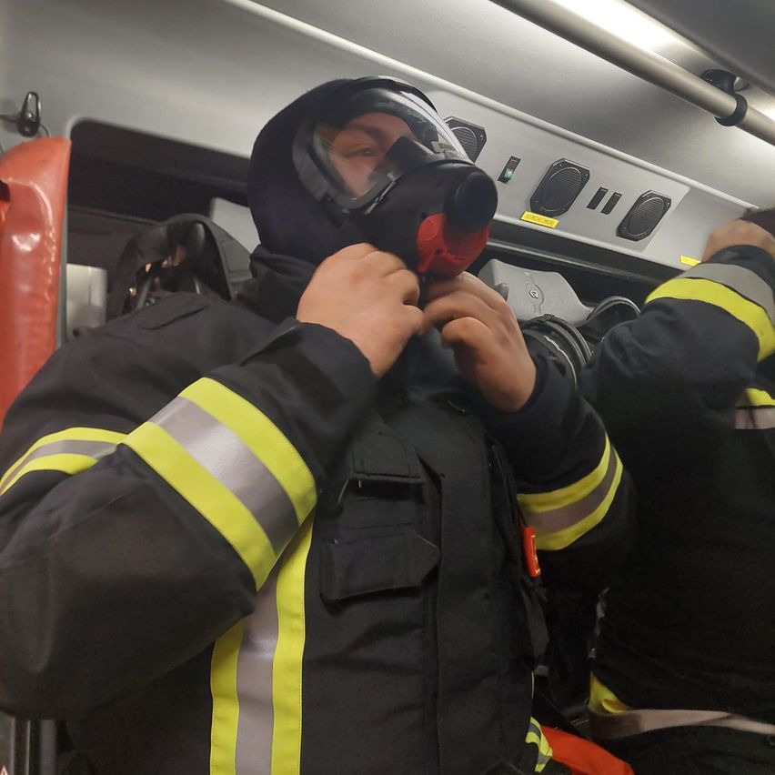 Ein Feuerwehrmann justiert seine Maske in einem Fahrzeug, wobei ein Sicherheitsweste und ein Helm im Hintergrund sichtbar sind.