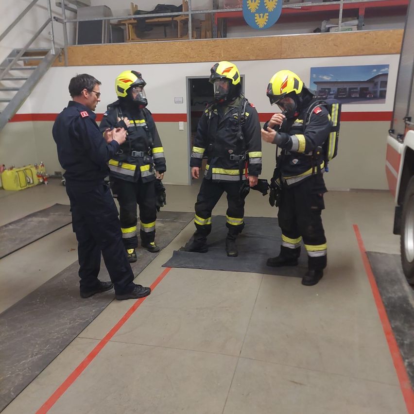 Drei Feuerwehrleute stehen in einem Übungsbereich mit einem Mann in Uniform. Sie tragen gelbe Helme und schwarze Uniformen mit gelben Streifen. Eine orange Linie ist auf dem Boden.