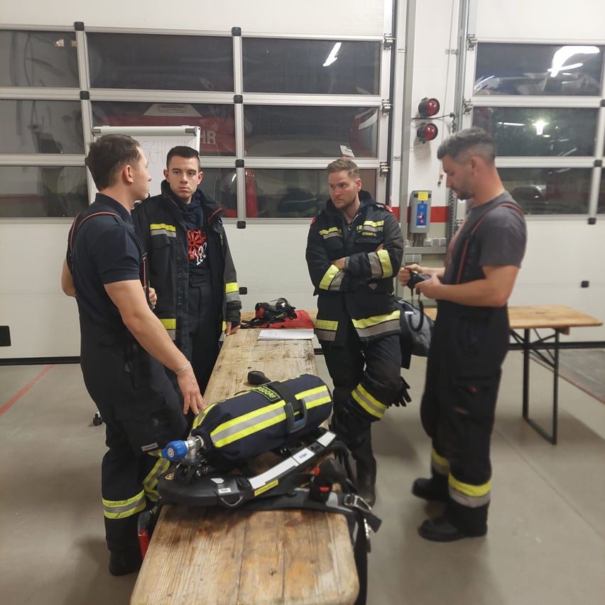 Vier Feuerwehrleute stehen in einem Raum, vermutlich einer Feuerwache. Sie tragen Uniformen und Helme, und einer hält ein Werkzeug. Sie scheinen etwas zu besprechen.