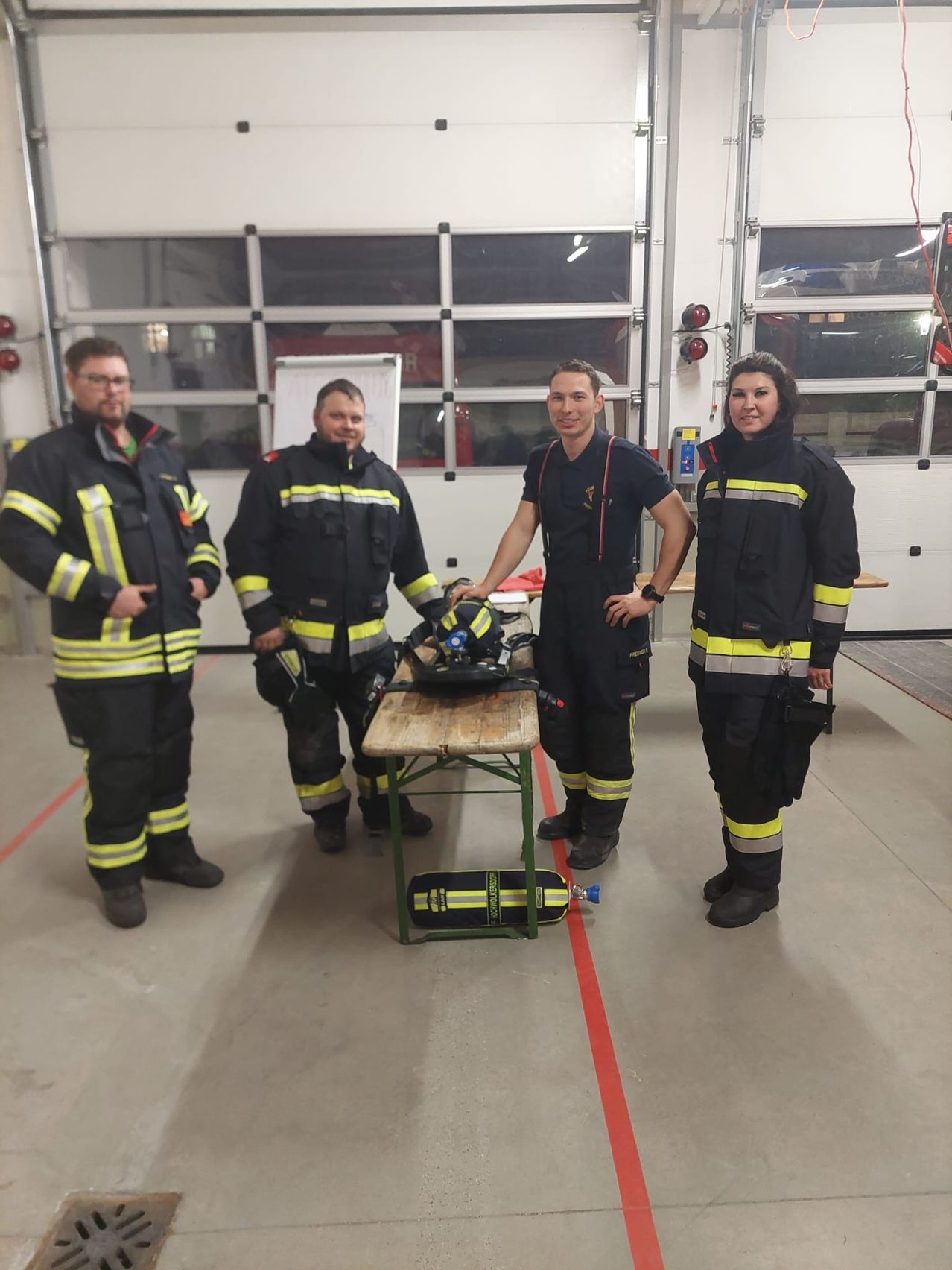 Vier Feuerwehrleute in Uniform stehen in einem Raum mit einem Tisch in der Mitte, wahrscheinlich in einer Feuerwache.
