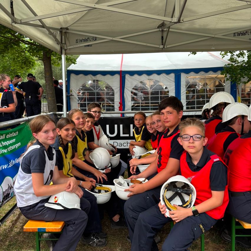 A group of children and young adults in safety gear and helmets sit under a tent with a banner and a crowd in the background.