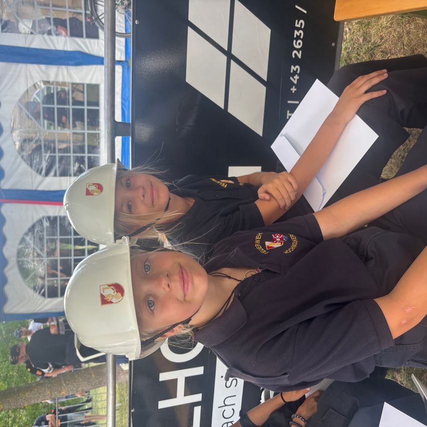 Two young girls in matching uniforms and white helmets pose for a photo in an outdoor setting. Behind them is a black banner with white squares and numbers.