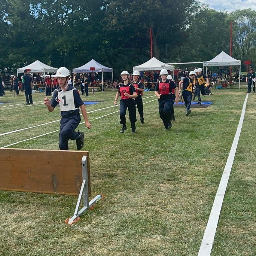 A group of people wearing helmets and numbered shirts are running on a field with hurdles and tents in the background.