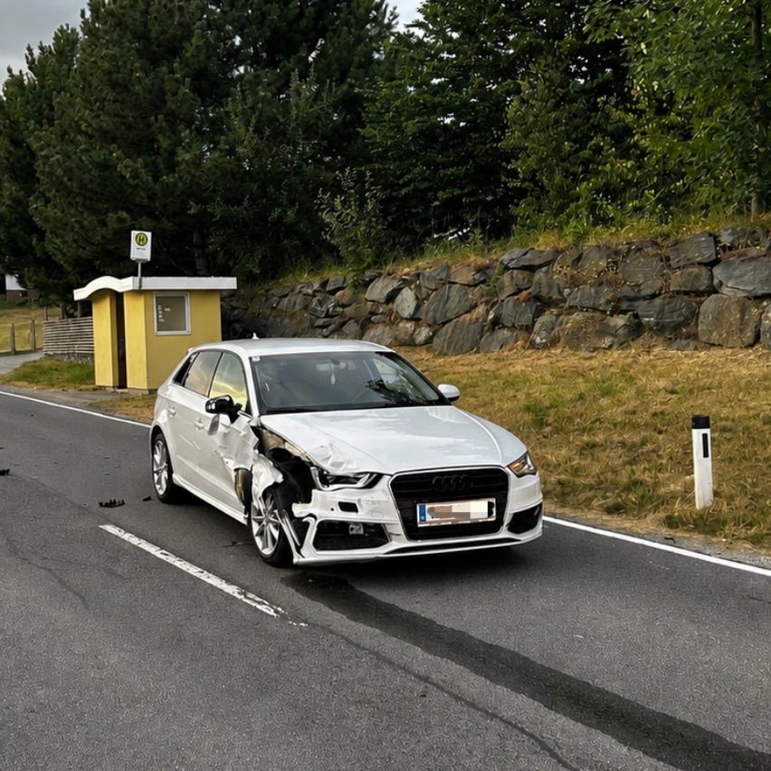 A silver Audi A3 with front-end damage is parked on the side of the road. The car has a license plate and is near a stone wall.