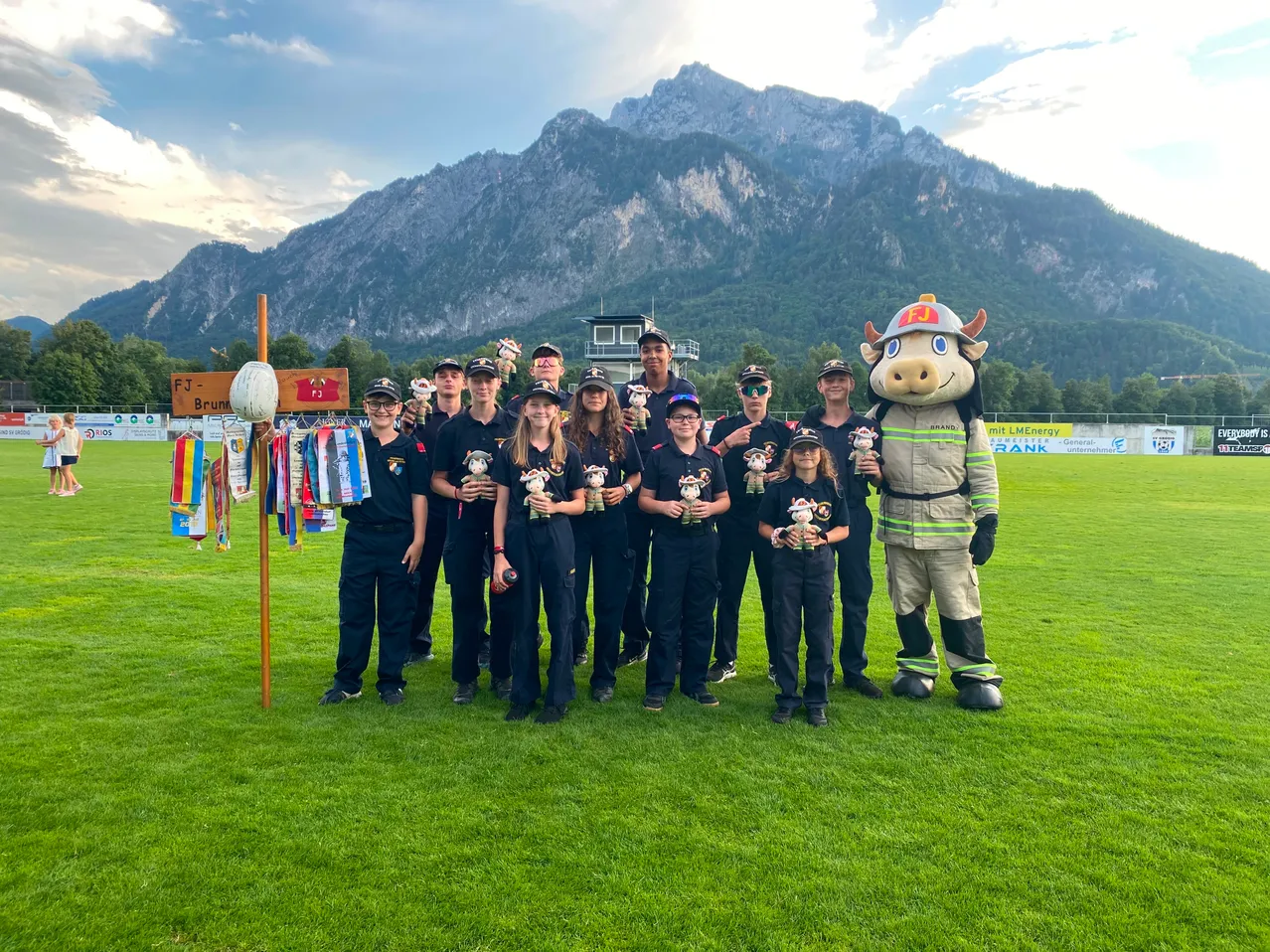 Eine Gruppe von Menschen in schwarzen Uniformen und Mützen posiert für ein Foto auf einem Rasenfeld mit Bergen im Hintergrund. Ein Maskottchen in einer Feuerwehruniform steht unter ihnen.