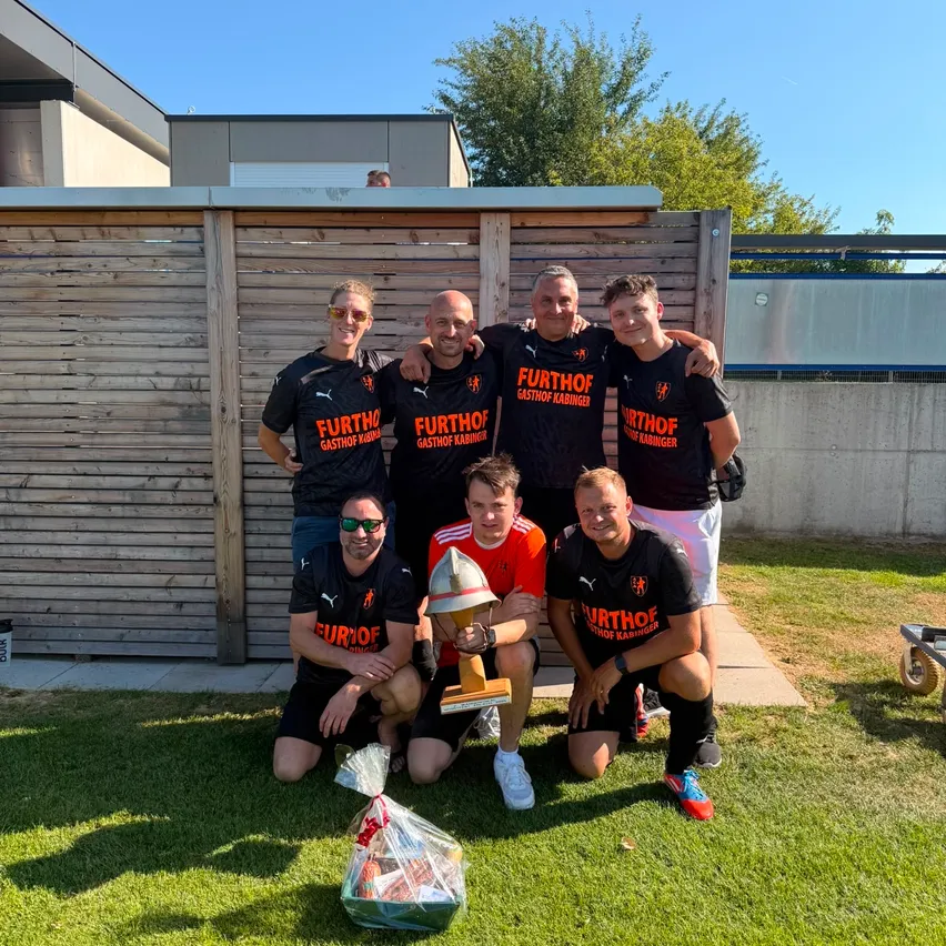 Eine Gruppe von Männern in schwarzen Trikots mit orangefarbenem Schriftzug posiert mit einer Trophäe auf einem Rasenfeld.