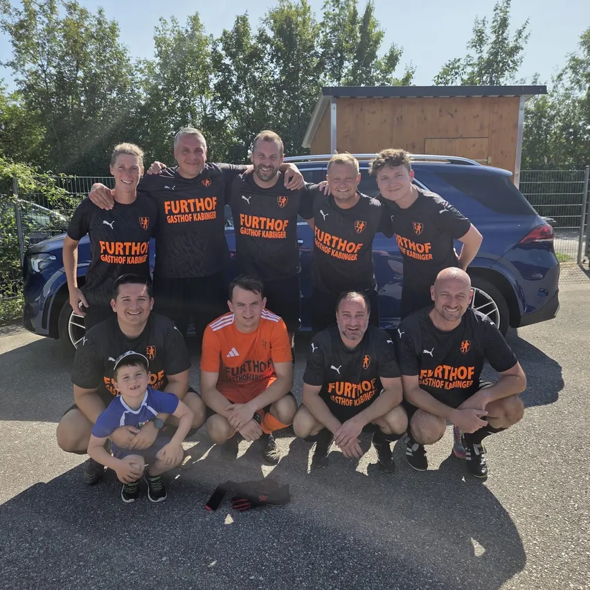 Eine Gruppe von Männern und einem Jungen in schwarzen und orangefarbenen Trikots mit dem Wort Furthof darauf posiert für ein Foto vor einem blauen Auto.