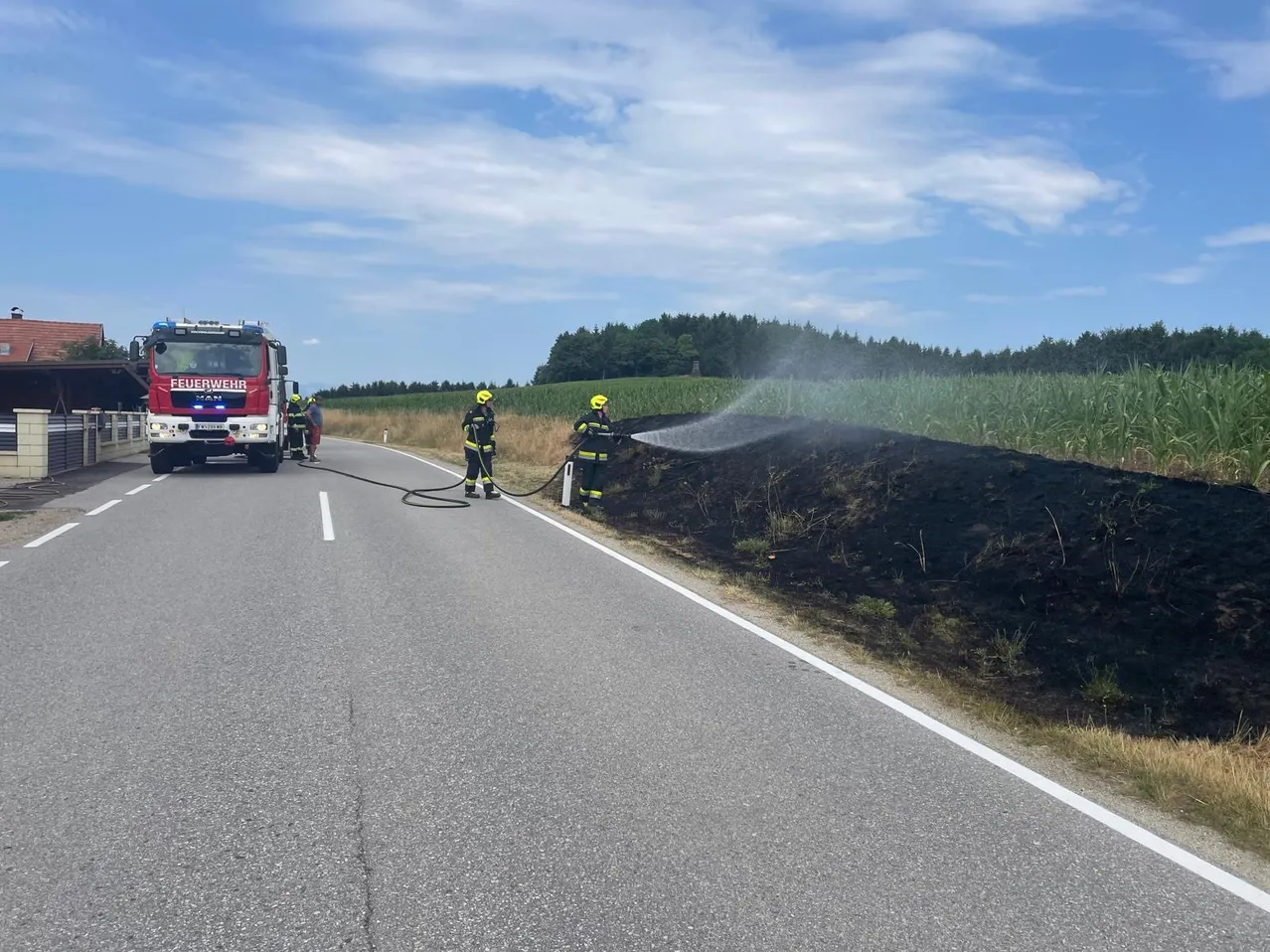 Zwei Feuerwehrleute sprühen Wasser auf einen brennenden Erdhaufen am Straßenrand, ein Feuerwehrauto ist in der Nähe.