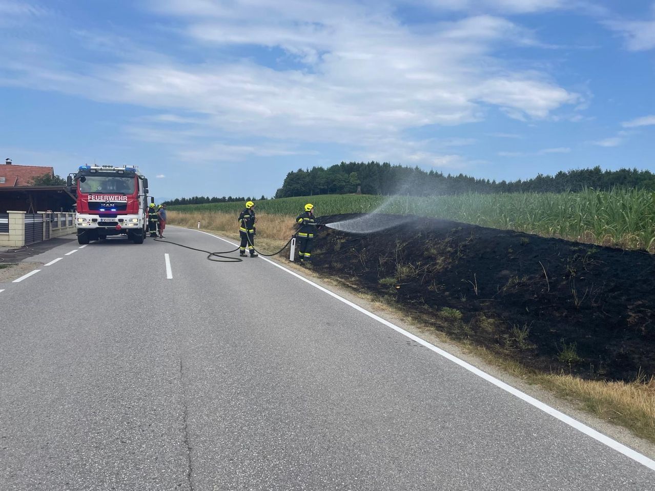 Zwei Feuerwehrleute sprühen Wasser auf einen brennenden Erdhaufen am Straßenrand, ein Feuerwehrauto ist in der Nähe.