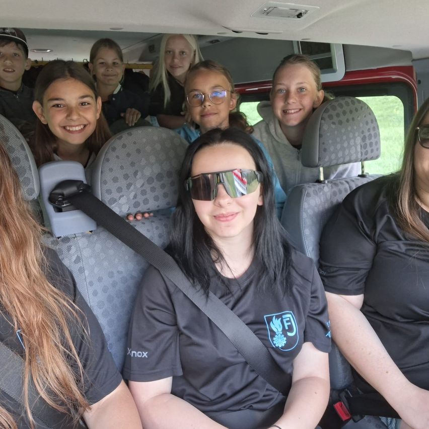 Eine Gruppe junger Mädchen und Frauen in einem Fahrzeug, alle tragen schwarze T-Shirts mit einem Logo. Das Mädchen vorne trägt eine Sonnenbrille und einen Sicherheitsgurt.