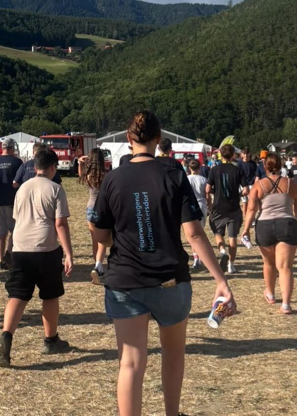 Eine Gruppe von Menschen läuft auf einem Erdfeld mit einem bewaldeten Hügel im Hintergrund. Einige tragen Freizeitkleidung, während andere Uniformen tragen. Eine Frau vorne trägt ein schwarzes Shirt mit blauer Schrift.