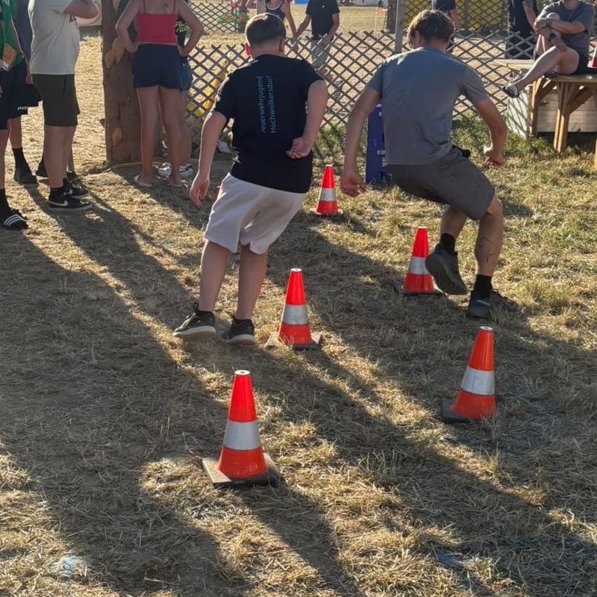 Mehrere Personen nehmen an einem Geschicklichkeitsparcours mit orangefarbenen Verkehrskegeln auf einem Grasfeld teil.