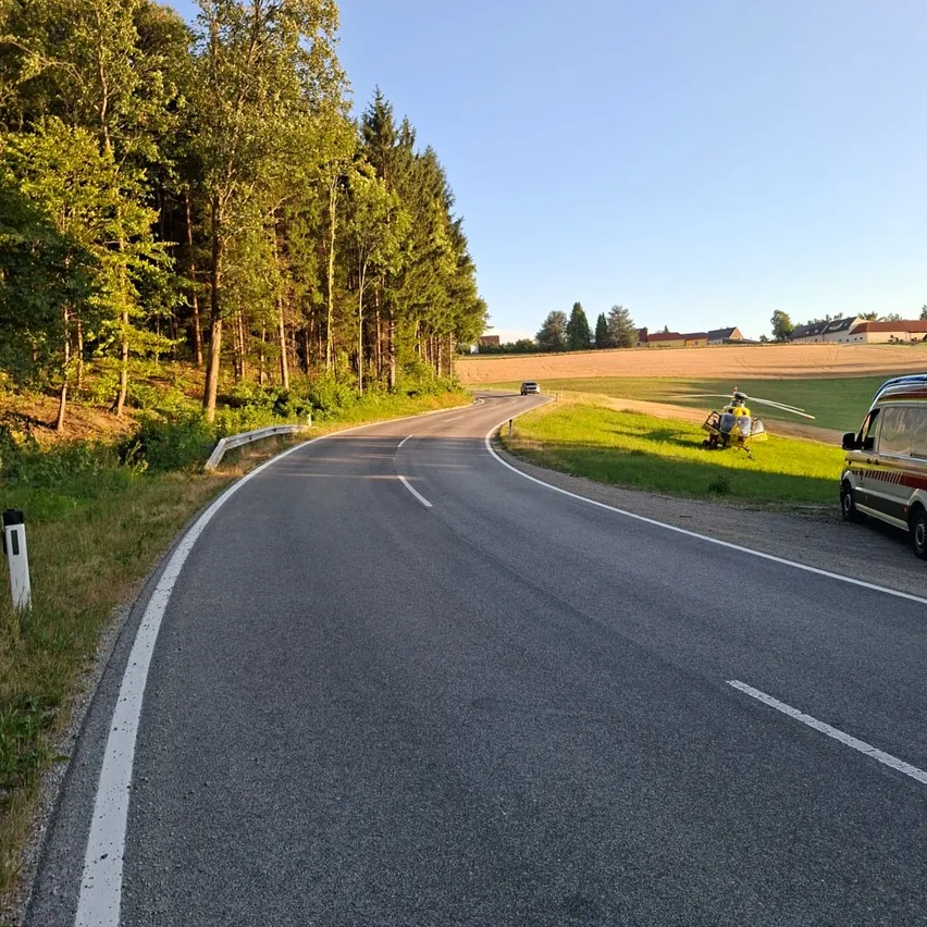 Eine leere Straße mit einem Hubschrauber am Grasrand, umgeben von Bäumen und einem entfernten Feld.