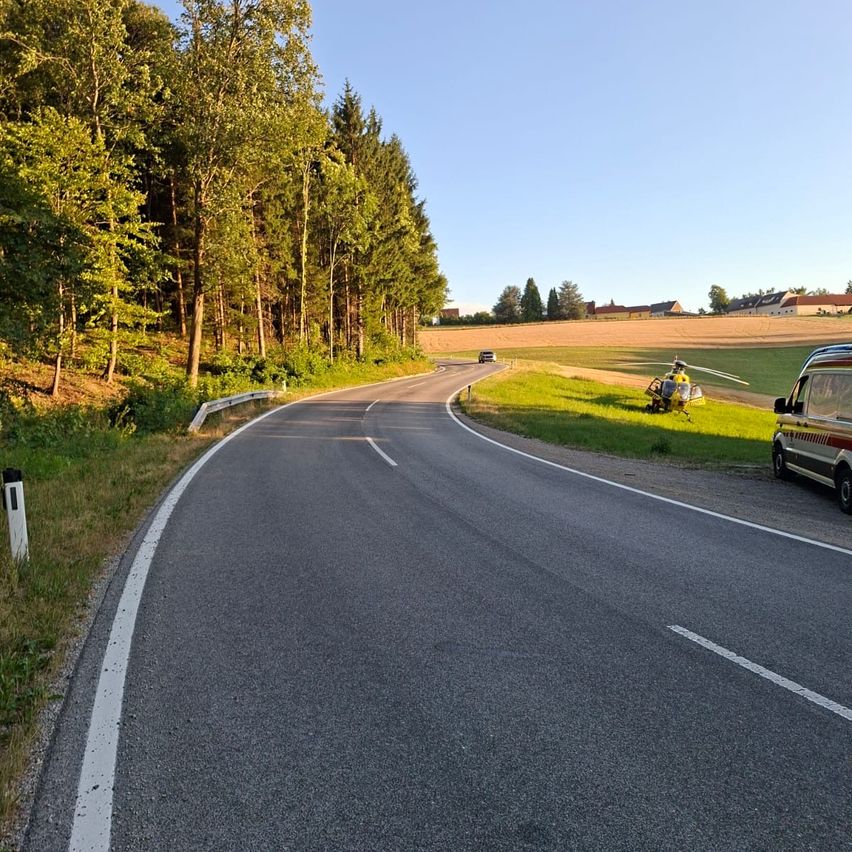 Eine leere Straße mit einem Hubschrauber am Grasrand, umgeben von Bäumen und einem entfernten Feld.