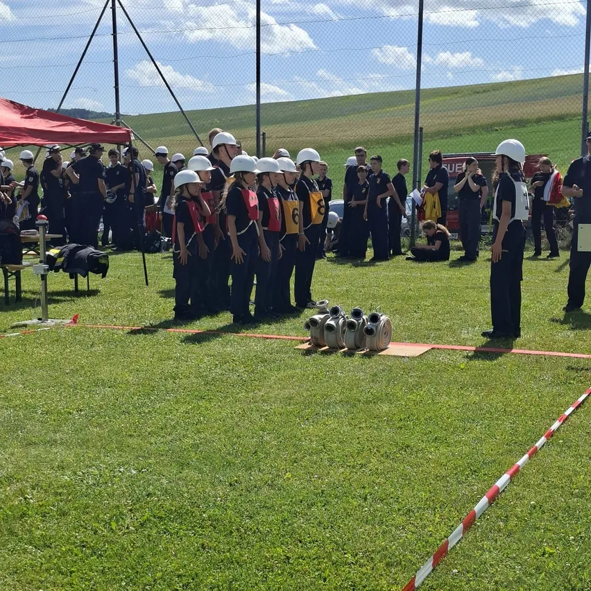 Eine Gruppe von Menschen in Helmen und Uniformen steht auf einem Feld mit Ausrüstung vor ihnen. Dahinter stehen weitere Menschen, und einige sitzen unter einem roten Zelt. In der Ferne ein Auto und ein Zaun.