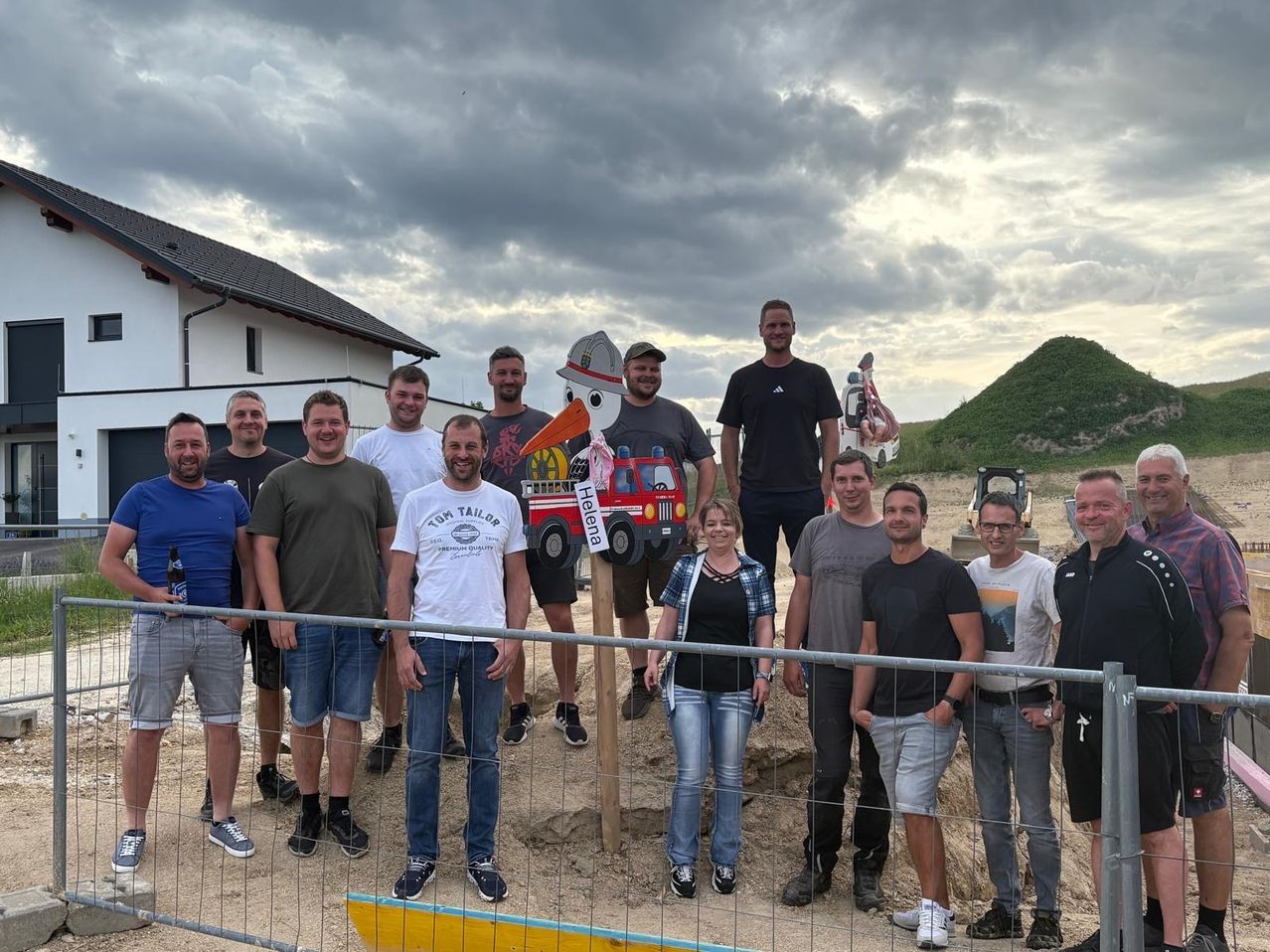 Eine Gruppe von Menschen posiert für ein Foto an einer Baustelle, mit einem Maskottchen im Hintergrund. Sie stehen hinter einem Zaun mit einem Haus und einem bewölkten Himmel darüber.