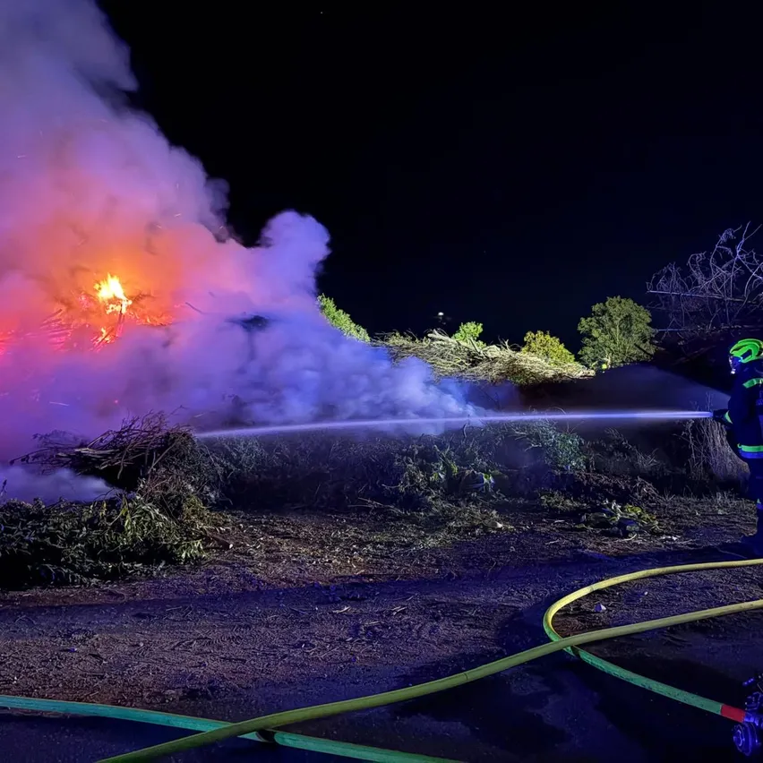 Zwei Feuerwehrleute in voller Ausrüstung benutzen einen Schlauch, um ein Feuer in der Nacht zu löschen. Das Feuer brennt einen Holzhaufen und ist von Rauch umgeben.