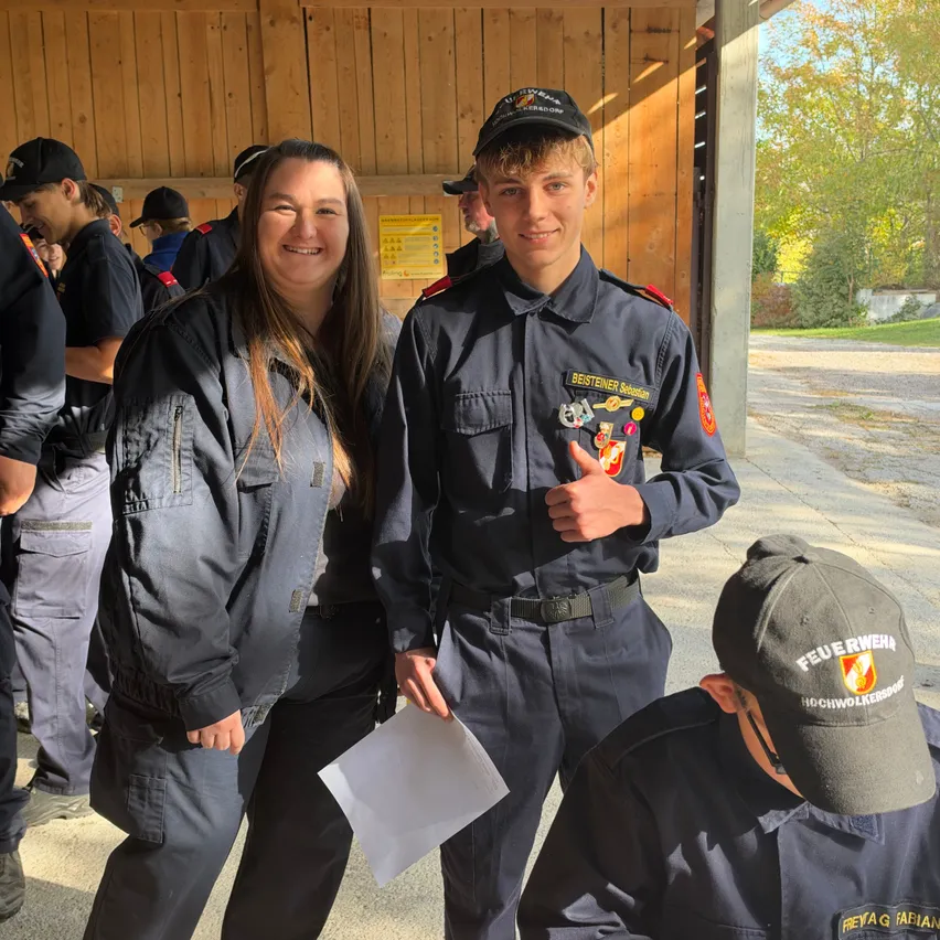 Eine Frau und ein junger Mann in Uniformen stehen zusammen und lächeln, mit einem jungen Jungen in Uniform vor ihnen. Sie sind draußen, mit einer Holzstruktur und Bäumen im Hintergrund.