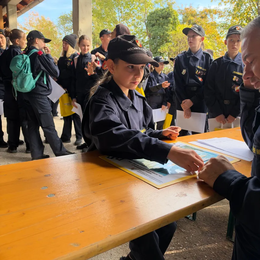 Ein junges Mädchen in Uniform sitzt an einem Tisch und schreibt auf ein Stück Papier, während ein Mann in Uniform neben ihr steht. Dahinter stehen eine Gruppe uniformierter Schüler in einer Reihe, wahrscheinlich warten sie auf ihren Zug.
