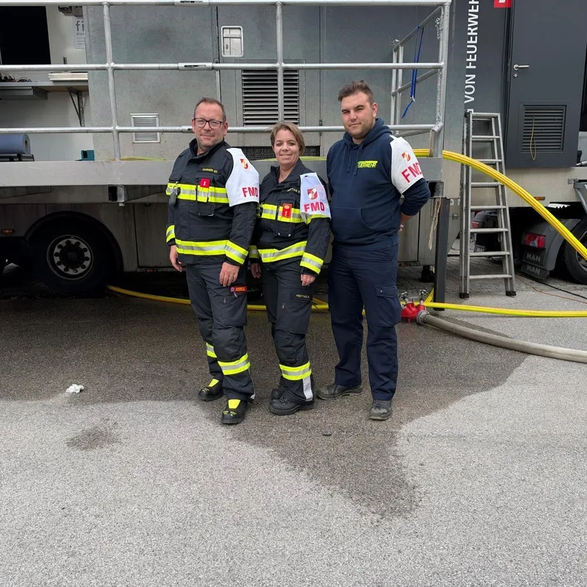 Drei Feuerwehrleute stehen vor einem LKW, zwei von ihnen tragen reflektierende Kleidung.