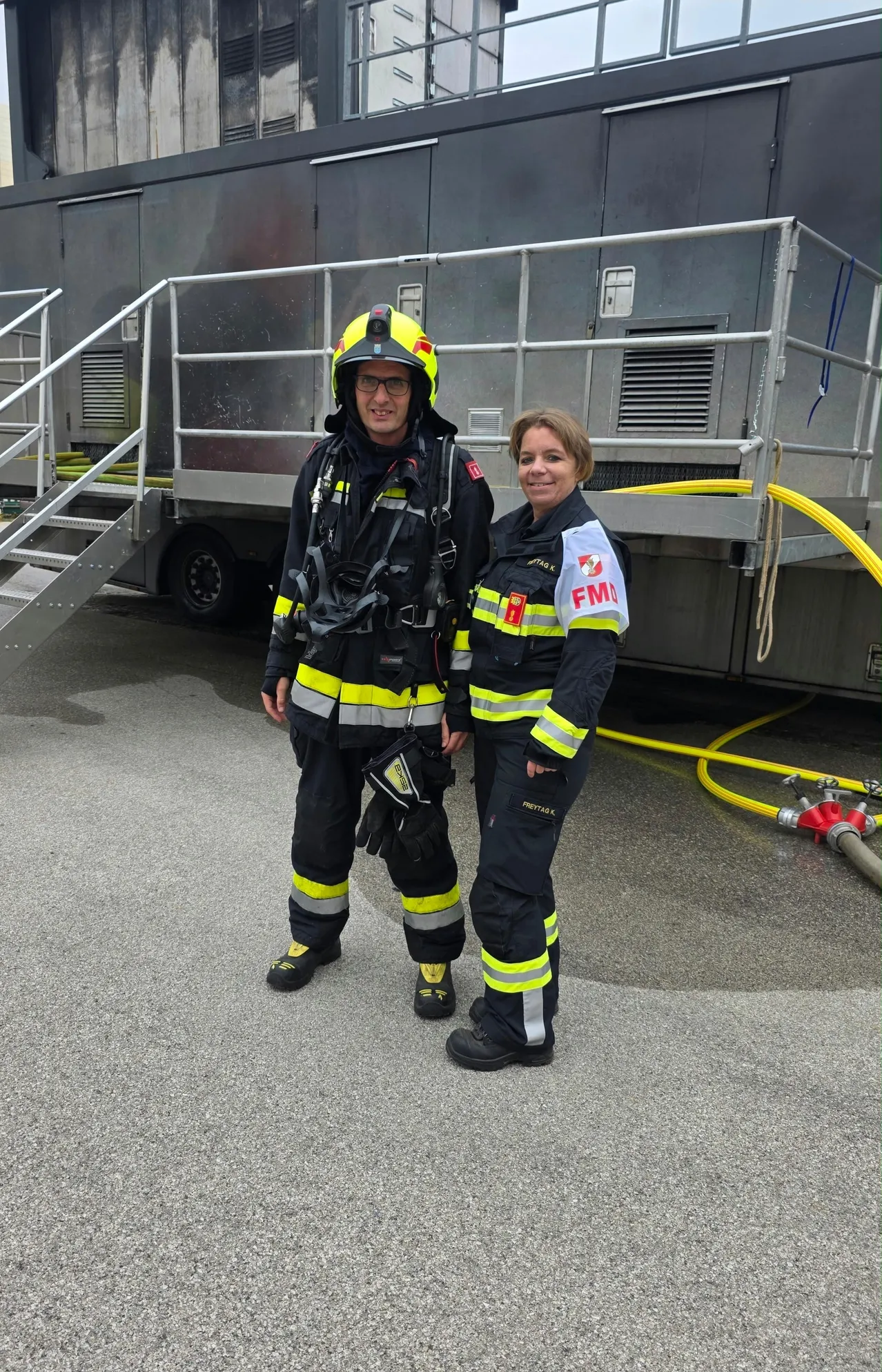 Zwei Feuerwehrleute, ein Mann und eine Frau, stehen vor einem Feuerwehrwagen. Der Mann trägt einen Helm und eine Maske, während die Frau eine reflektierende Weste trägt. Sie sind beide in voller Feuerwehrausrüstung.