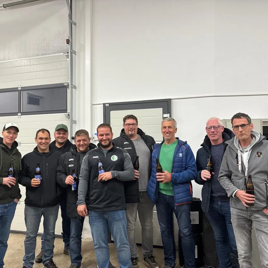 Eine Gruppe von Männern steht in einem Raum, lächelt und posiert für ein Foto. Sie halten alle Flaschen Bier in den Händen.